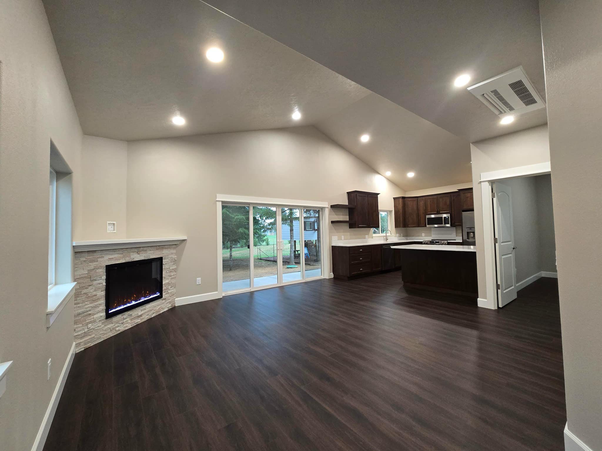 Open-concept home interior with modern kitchen, dark wood floors, stone fireplace, and sliding glass doors to backyard.